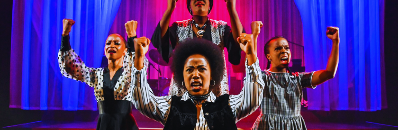 Four performers stand on stage with raised fists and intense expressions, set against a backdrop of dramatic pink and blue lighting and curtains, creating a powerful and dynamic scene.