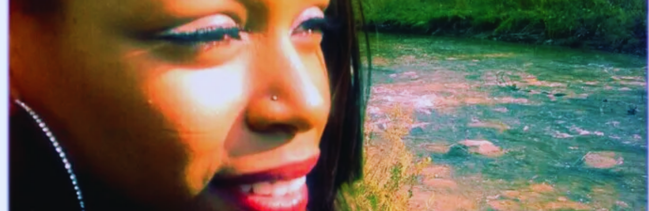 A person with dark hair smiles near a flowing creek, with sunlight highlighting their face. Green grass lines the creek in the background.